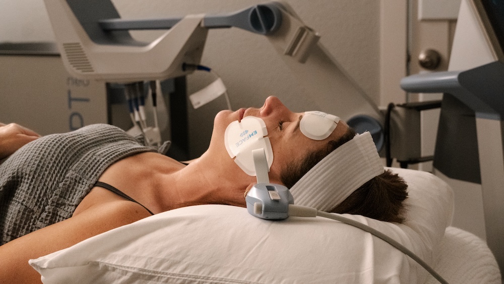 Photo of woman laying in treatment room with Emface applicators on her cheeks for facial contouring in Waterloo